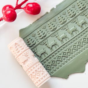 May include: A light pink polymer clay roller with a detailed winter sweater pattern, including reindeer and trees, next to a green clay sheet with the same design. Red berries on a branch are in the background.