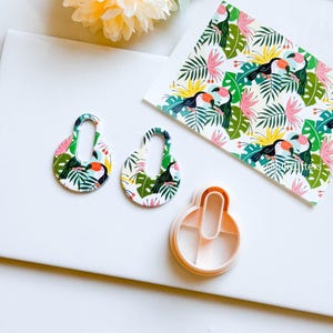 May include: Two white, oval-shaped earrings with a tropical toucan and leaf pattern. The earrings are next to a white plastic cookie cutter in the shape of the earrings.