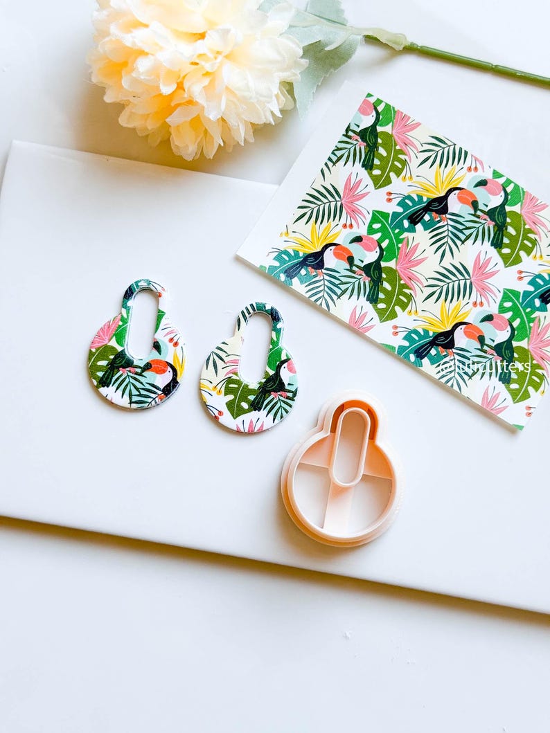 May include: Two white, oval-shaped earrings with a tropical toucan and leaf pattern. The earrings are next to a white plastic cookie cutter in the shape of the earrings.