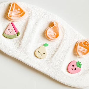 May include: Five pink plastic cookie cutters shaped like a watermelon, pear, strawberry, and two different types of pears. The cutters have cute cartoon faces on them.