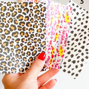 Op de afbeelding: Een hand houdt drie papieren met patronen vast. De eerste heeft een luipaardprint in zwart, bruin en bruin. De tweede heeft een roze, gele en rode luipaardprint. De derde heeft zwarte stippen op een witte achtergrond.