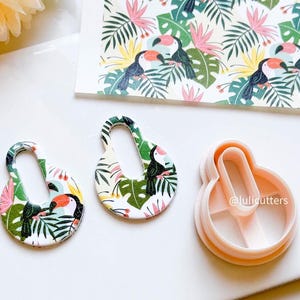 May include: Two white, oval-shaped cookie cutters with a tropical toucan and leaf design. The cutters are on a white surface next to a sheet of paper with a repeating pattern of toucans and leaves. The paper is labelled "@lulicutters".