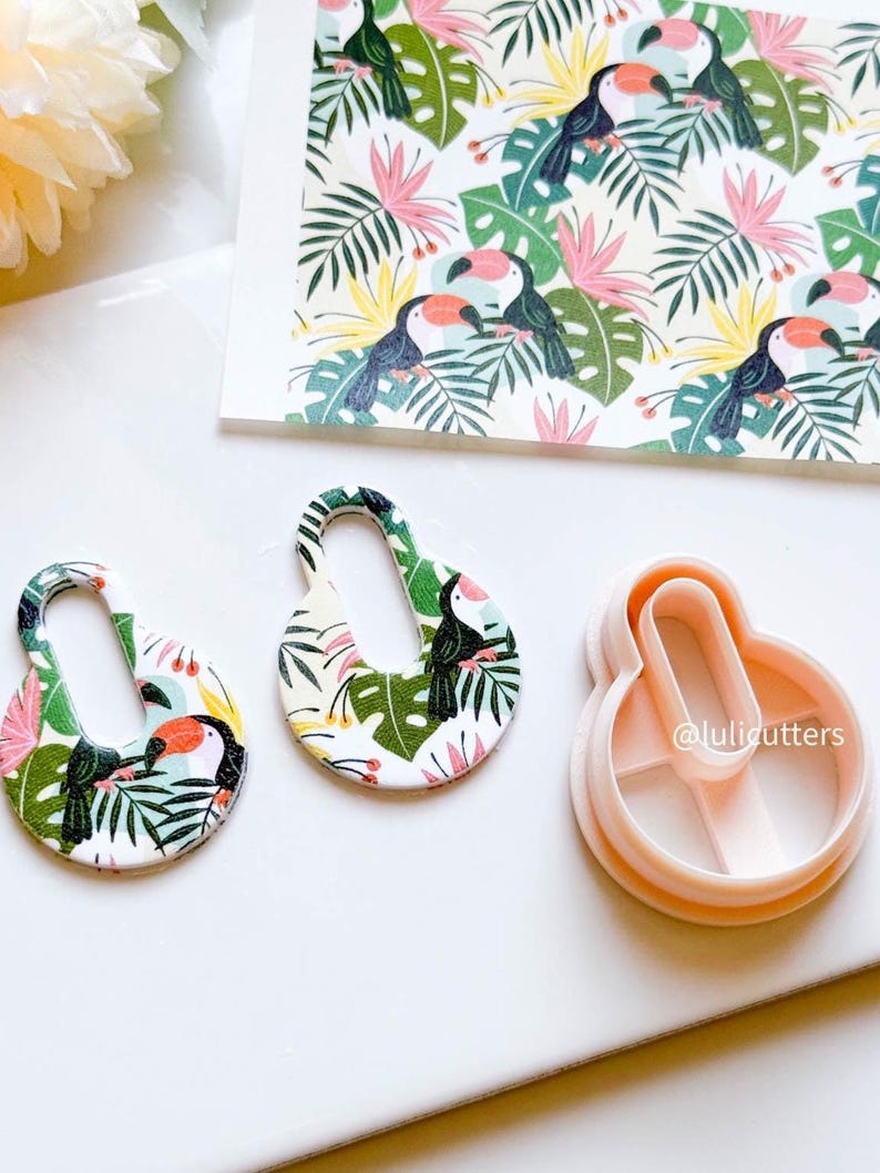 May include: Two white, oval-shaped cookie cutters with a tropical toucan and leaf design. The cutters are on a white surface next to a sheet of paper with a repeating pattern of toucans and leaves. The paper is labelled "@lulicutters".