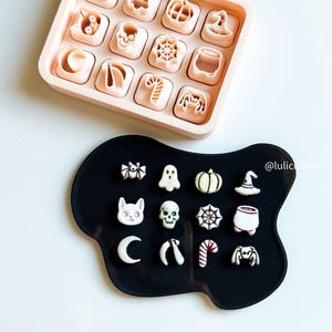 May include: A collection of Halloween-themed cookie cutters in a pink tray and on a black surface. The cutters include shapes like a spider, ghost, pumpkin, witch hat, skull, and candy cane. The cutters are in a variety of sizes and are made of plastic.