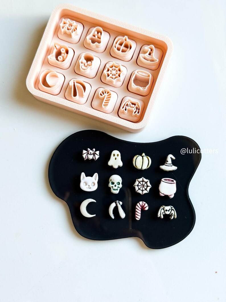 May include: A collection of Halloween-themed cookie cutters in a pink tray and on a black surface. The cutters include shapes like a spider, ghost, pumpkin, witch hat, skull, and candy cane. The cutters are in a variety of sizes and are made of plastic.