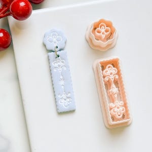 May include: A light blue earring with a floral design and a gold-colored hook, next to two cookie cutters. One cookie cutter is a flower shape, and the other is a rectangular shape with a floral design. The items are on a white surface.
