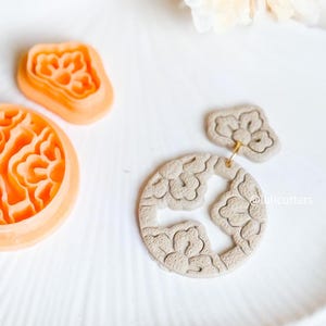 May include: A set of three polymer clay cutters featuring a floral design. The cutters are stacked on top of each other, with the largest cutter on the bottom and the smallest cutter on top. The cutters are a light orange color.