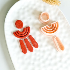Könnte beinhalten: Eine Sammlung von Ausstechern aus terrakottafarbenem Polymer Clay in verschiedenen geometrischen Formen, darunter ein Kreis, ein Halbkreis und längliche Formen. Die Ausstecher sind auf einem weißen, strukturierten Keramikteller angeordnet, mit getrockneten floralen Akzenten im Hintergrund.