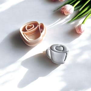 May include: Two cookie cutters shaped like coffee cups, one beige and one silver, each with a heart design. Pink tulips with green stems and leaves are in the background, all on a white surface.