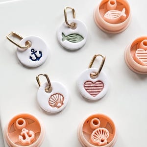 May include: White ceramic earrings with gold hoops, featuring nautical designs like an anchor, fish, seashell, and heart. Matching pink molds with the same designs are also present, suggesting a crafting or jewelry-making theme.