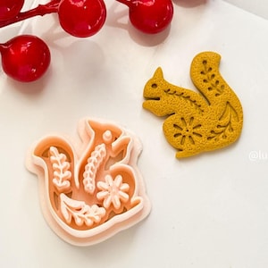 May include: A light orange squirrel-shaped cookie cutter next to a yellow squirrel-shaped cookie. The cookie cutter has floral and leaf details. A red berry branch is in the background.