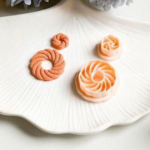 Könnte beinhalten: Draufsicht auf vier orangefarbene und pfirsichfarbene Ton-Ausstecher in verschiedenen Spiral- und Kreisformen. Die Ausstecher liegen auf einem weißen Teller mit gewelltem Rand, im Hintergrund eine hellblaue Blume.