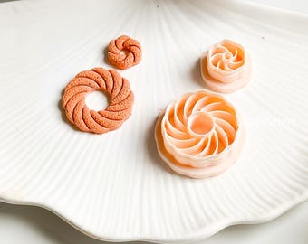 May include: An overhead shot displays four orange and peach-colored clay cutters in various spiral and circular shapes. The cutters are arranged on a white, scalloped-edge plate, with a soft blue flower in the background.