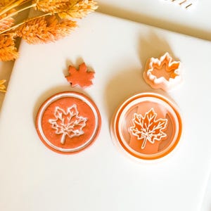 May include: Orange maple leaf cookie cutters and stamps. The set includes a small leaf, a leaf-shaped cutter, and two round stamps with leaf designs. The items are arranged on a white surface with orange foliage.