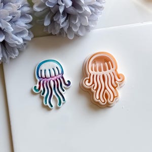 Op de afbeelding: Twee koekjessnijders in de vorm van een kwal. Eén is een afgewerkt product met een wit, blauwgroen, paars en blauw ontwerp. De andere is een onafgewerkte, perzikkleurige snijder. De snijders liggen op een wit oppervlak met een bloemig accent.