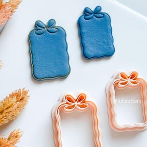 Könnte beinhalten: Zwei blaue, geschenkförmige Ausstecher mit goldenem Rand und Schleifendetail. Zwei rosa, geschenkförmige Ausstecher mit orangefarbenen Schleifendetails sind ebenfalls abgebildet. Die Ausstecher befinden sich auf einer weißen Oberfläche.