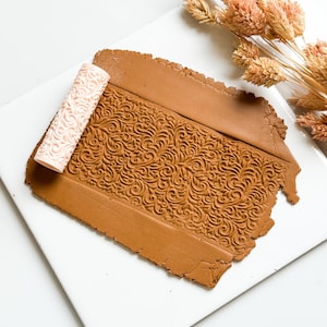 Könnte beinhalten: Eine hellbraune Tonplatte mit geprägtem floralem Muster, neben einer hellrosa gemusterten Rolle. Der Ton liegt auf einer weißen Oberfläche, mit getrockneten Blumen im Hintergrund. Bastelbedarf.