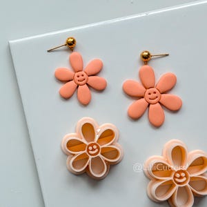 May include: Peach-colored daisy earrings with gold-colored posts and a smiling face in the center of each flower. Two daisy-shaped cookie cutters with smiling faces are also visible.