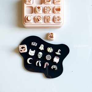 May include: A collection of Halloween-themed cookie cutters in a variety of shapes, including pumpkins, ghosts, and spiders. The cutters are arranged on a black surface, with a pink storage tray above. The image includes the text "@lulicutters".