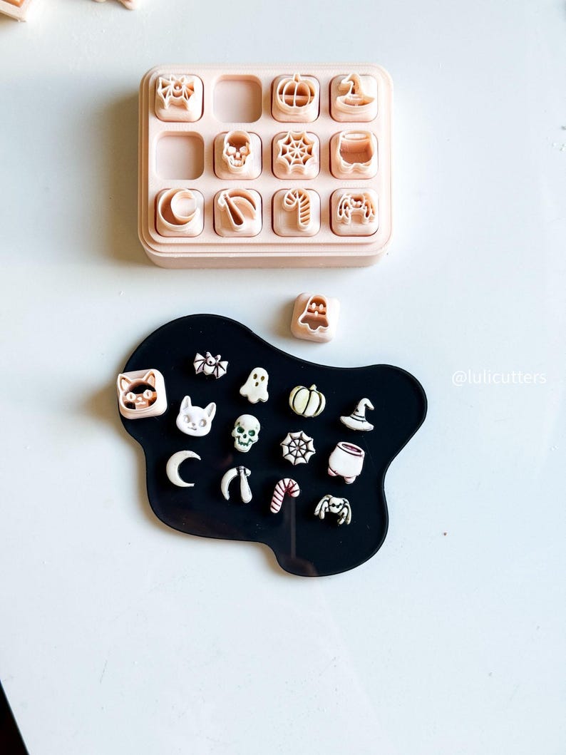 May include: A collection of Halloween-themed cookie cutters in a variety of shapes, including pumpkins, ghosts, and spiders. The cutters are arranged on a black surface, with a pink storage tray above. The image includes the text "@lulicutters".