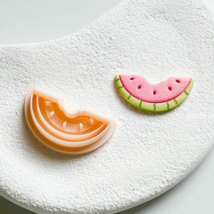 May include: Two watermelon-themed cookie cutters. One is a light orange colour, shaped like a slice with a bite taken out. The other is a pink watermelon slice with green rind and black seed details. Both are on a white textured surface.