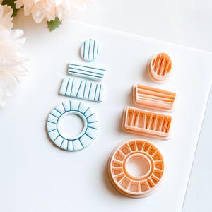 Könnte beinhalten: Draufsicht auf Polymer Clay Cutter in verschiedenen Formen und Größen. Die Cutter sind in Blau- und Pfirsichtönen gehalten. Die blauen Cutter haben dünne, dunkelblaue Linien. Die Pfirsich-Cutter haben dünne, vertikale Linien. Die Cutter sind auf einer weißen Oberfläche angeordnet.