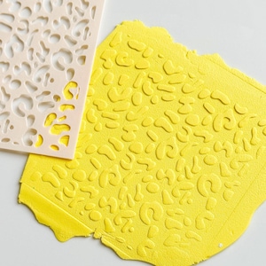 May include: A light beige stencil with an organic pattern is placed over a textured yellow surface. The yellow surface has a raised pattern that mirrors the stencil design. The image is a close-up shot, highlighting the textures and colours.