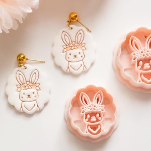 Può includere: Orecchini bianchi e dorati e formine rosa. Gli orecchini presentano un design a coniglietto con una corona floreale. Le formine sono rosa e presentano anche un design a coniglietto con una corona floreale. Gli orecchini hanno perni dorati.