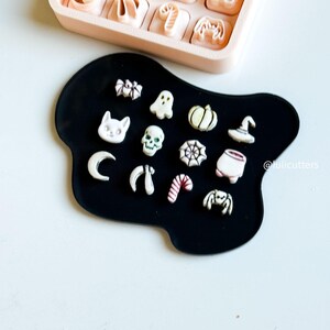 May include: A collection of Halloween-themed cookie cutters and shapes, including a cat, ghost, pumpkin, spider, and skull. The cutters are in a pink tray, and the shapes are on a black surface. The image has a light background.