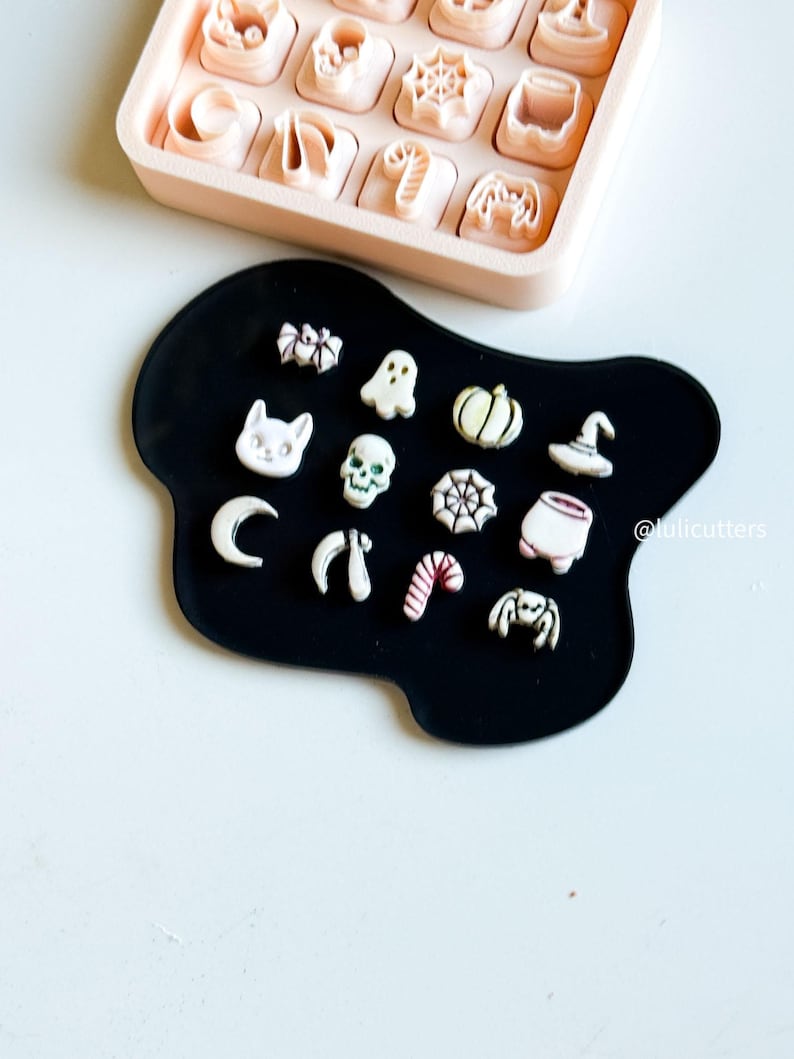 May include: A collection of Halloween-themed cookie cutters and shapes, including a cat, ghost, pumpkin, spider, and skull. The cutters are in a pink tray, and the shapes are on a black surface. The image has a light background.