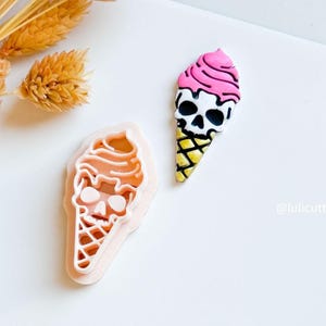 May include: A pink and white skull-shaped ice cream cone with a yellow waffle cone. Beside it is a light pink cookie cutter in the shape of the ice cream cone. The image is on a white background.