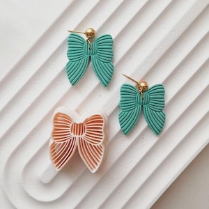 Op de afbeelding: Twee paar groene vlinder-oorbellen met gouden accenten. De oorbellen liggen op een wit oppervlak met een textuurpatroon.