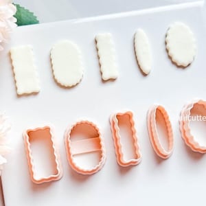 May include: A collection of cookie cutters in various shapes, including rectangles, ovals, and rounded rectangles. The cutters are light pink and white, with scalloped edges on some. The shapes are arranged on a white surface.