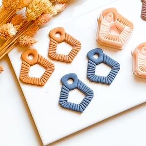 May include: A selection of geometric cookie cutters in shades of orange and blue are arranged on a white surface. The cutters feature a textured pattern and a hole at the top. Dried orange flowers are in the background.