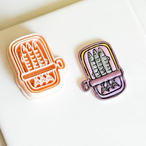 Op de afbeelding: Twee koekjessnijders met sardinethema. De ene is een lichtoranje plastic snijder en de andere is een beschilderde sardineblik met een gele rand, roze binnenkant en drie vissen. De snijders liggen op een wit oppervlak.