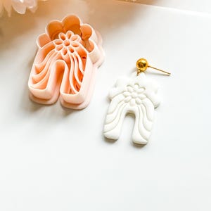 Op de afbeelding: Een lichtperzik kleurige polymeerklei cutter en een witte polymeerklei oorbel met een bloemenontwerp. De oorbel heeft een goudkleurige stud. De cutter heeft de vorm van de oorbel. De oorbel heeft een gebogen, boogachtige vorm.
