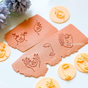 May include: A set of six cookie cutters featuring different abstract face designs. The cutters are made of a light orange plastic and are arranged on a white surface. The cutters are perfect for creating unique and modern cookies.