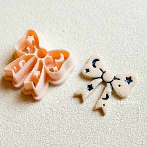 Op de afbeelding: Twee koekjessnijders in de vorm van een strik. Eén is licht perzik van kleur en de andere is beige met zwarte maan- en sterdetails. De perziksnijder heeft uitgesneden vormen van manen en sterren. De beige strik heeft een geschilderd ontwerp van manen en sterren.