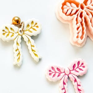 Pode incluir: Uma vista aérea mostra três ornamentos em forma de laço. Um é branco com detalhes dourados e uma corrente de bolinhas dourada. Os outros dois são rosa, com diferentes tons de rosa. Decorativos.