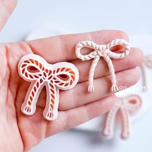 May include: Two bow-shaped cookie cutters are shown. One is white with orange detailing, the other is a solid off-white colour. The cutters feature a rope-like design, ideal for decorative cookies or fondant accents.