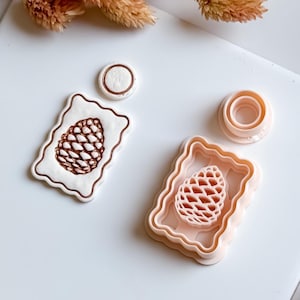May include: A set of light pink and white cookie cutters in the shape of a pine cone. The set includes a pine cone shaped cutter, a rectangular frame cutter, and a small circle cutter. The pine cone design is detailed and textured.