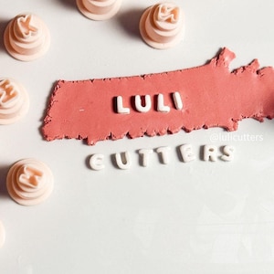 以下が含まれることがあります： 文字の形をした薄いピンク色の積層カッターが、白い文字で「LULI CUTTERS」と書かれた赤い粘土の周りに散らばっています。カッターは白い表面にあります。