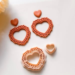 Peut inclure: Ensemble de découpoirs à biscuits en forme de cœur et de petits biscuits en forme de cœur. Les découpoirs sont dans des tons de rose et d'orange, avec un bord festonné décoratif. Les biscuits sont d'une couleur similaire. Sur une surface blanche.