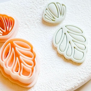 May include: Close-up of several leaf-shaped cookie cutters. Two are light orange, and two are light green. The cutters are on a white surface, dusted with a fine white powder. The cutters are designed to create leaf-shaped cookies.
