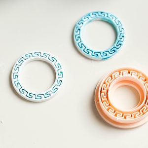 May include: Three decorative rings with a Greek key design. One ring is white with teal accents, another is light blue with darker blue accents, and the third is peach-colored. The rings are on a white surface.