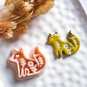 May include: A fox-shaped cookie cutter and a decorative fox figurine. The cookie cutter is light orange with a white outline and floral details. The figurine is yellow with gold accents and floral patterns. Dried wheat stalks are in the background.