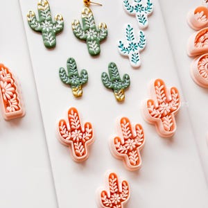 Könnte beinhalten: Eine Sammlung von Ohrringen und dekorativen Elementen in Kaktusform. Die Ohrringe sind in Grün, Weiß und Orange gehalten und mit goldenen Akzenten versehen. Die Kaktus-Designs zeigen Blumen- und Blättermuster auf weißem Hintergrund.