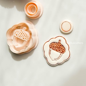May include: A set of light pink acorn-shaped cookie cutters and a stamped biscuit. The cutters feature a detailed acorn design and a scalloped edge. The stamped biscuit has a detailed acorn design with a brown top and white base.