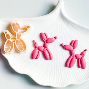 Pode incluir: Três cortadores de biscoitos em forma de cão de balão rosa em um prato branco. Um cortador de biscoitos tem a forma de um cão de balão, e os outros dois estão recheados com fondant rosa.