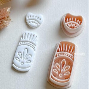 May include: Four white and peach-colored cookie cutters with decorative floral and geometric designs. The cutters are in various shapes, including a heart and a rectangular shape. The image also includes dried flowers in the background.